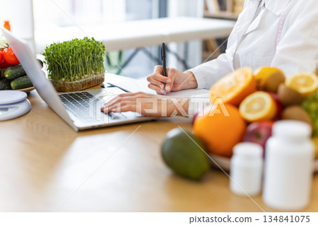 A dietitian works on a laptop and takes notes surrounded by fresh fruits, vegetables, and supplements, promoting healthy eating. A dietitian works on a laptop and takes notes surrounded by fresh fruits, vegetables, and supplements, promoting healthy eating. 134841075
