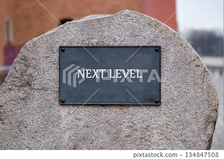 A stone monument displays a dark metallic plate with the words NEXT LEVEL in white lettering, set against a blurred backdrop of buildings in an urban setting A stone monument displays a dark metallic plate with the words NEXT LEVEL in white lettering, set against a blurred backdrop of buildings in an urban setting 134847508