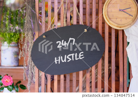A dark chalkboard sign that reads 24 7 HUSTLE hangs on a rustic wooden lattice wall. Beside it is a light wood clock and a bouquet of flowers, creating a warm, inviting atmosphere A dark chalkboard sign that reads 24 7 HUSTLE hangs on a rustic wooden lattice wall. Beside it is a light wood clock and a bouquet of flowers, creating a warm, inviting atmosphere 134847514