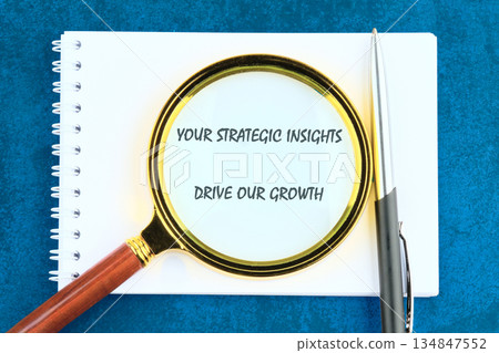 On a blue surface, a spiral notebook displays the phrase YOUR STRATEGIC INSIGHTS DRIVE OUR GROWTH under a magnifying glass next to a pen On a blue surface, a spiral notebook displays the phrase YOUR STRATEGIC INSIGHTS DRIVE OUR GROWTH under a magnifying glass next to a pen 134847552
