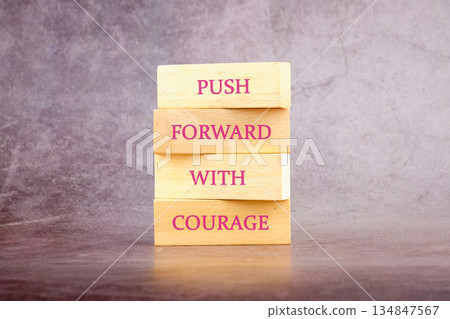 A stack of four wooden blocks display the words PUSH FORWARD WITH COURAGE. A motivational message in pink against a faded purple backdrop A stack of four wooden blocks display the words PUSH FORWARD WITH COURAGE. A motivational message in pink against a faded purple backdrop 134847567