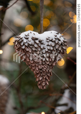 Handmade heart-shaped Christmas ornament made of natural seeds hangs on branches, covered with light snow, warm golden bokeh lights in the background 134849195