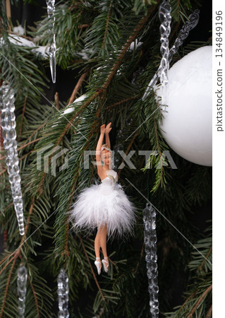 Ballerina ornament in white tutu hangs on green fir branches with crystal icicles, white bauble, creating an elegant festive winter close-up 134849196