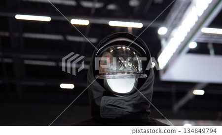 Visor reflection closeup capturing exhibition lights and ceiling architecture with glossy curvature and layered reflections on transparent shield, moody contrast and studio bokeh 134849794