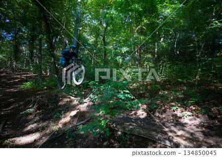 Electric mountain biker jumps a forest trail drop in lush green woods 134850045