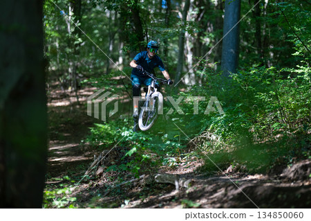 Mountain biker jumps over a trail obstacle in a forest on an electric mountain bike Mountain biker jumps over a trail obstacle in a forest on an electric mountain bike 134850060