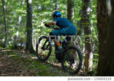 Rider on a forest trail: mountain biking in lush woods with helmet and gear Rider on a forest trail: mountain biking in lush woods with helmet and gear 134850129