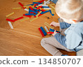 Child sitting on a wooden floor surrounded by colorful building toys, holding pieces and engaging in creative play. Warm home atmosphere emphasizes themes of early development, imagination and 134850707