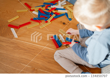 Child sitting on a wooden floor surrounded by colorful building toys, holding pieces and engaging in creative play. Warm home atmosphere emphasizes themes of early development, imagination and 134850707