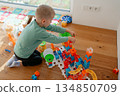 A young child sits on the floor playing with a colorful toy track set near a bright window. The scene captures creativity, learning and early childhood development in a warm home environment with 134850709