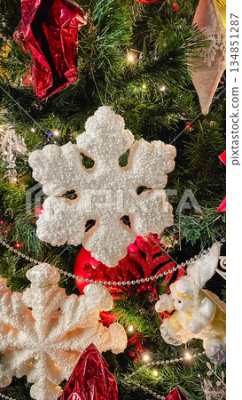 Close-up of a large white decorative snowflake hanging on a Christmas tree with red ornaments. Close-up of a large white decorative snowflake hanging on a Christmas tree with red ornaments. 134851287