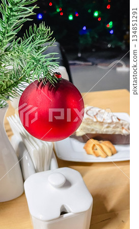 Close-up of a red matte Christmas ball on a spruce branch in a cafe setting with a blurred cake and cookies in the background. Close-up of a red matte Christmas ball on a spruce branch in a cafe setting with a blurred cake and cookies in the background. 134851291