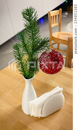 A red glitter Christmas ball on a spruce branch in a white vase on a wooden table in a cafe. 134851563