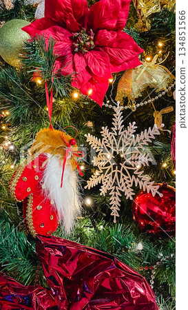 Close-up of a Christmas tree decorated with a red Santa mitten, a large poinsettia flower, a white snowflake, and warm fairy lights. Close-up of a Christmas tree decorated with a red Santa mitten, a large poinsettia flower, a white snowflake, and warm fairy lights. 134851566