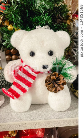 Close-up of a cute white decorative teddy bear wearing a red striped scarf and holding a pine cone, set against a Christmas tree background. Close-up of a cute white decorative teddy bear wearing a red striped scarf and holding a pine cone, set against a Christmas tree background. 134851569