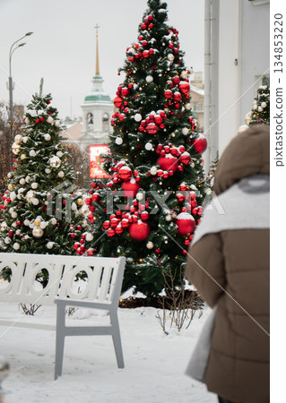 Decorated Christmas trees with red and white ornaments stand in snowy urban setting near white bench, festive winter atmosphere with holiday lights 134853220