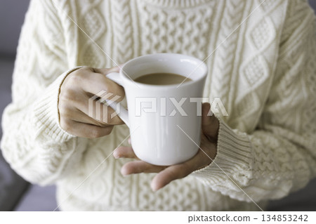 The hands of a person wearing a knitted dress holding a white mug of cafe au lait in both hands The hands of a person wearing a knitted dress holding a white mug of cafe au lait in both hands 134853242