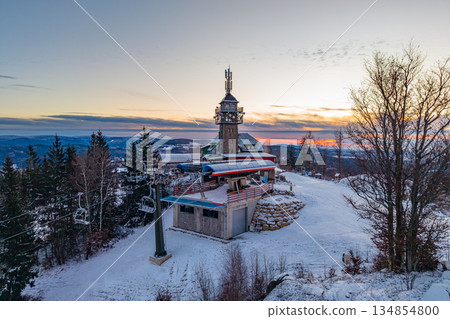 Tanvaldsky Spicak Ski Resort shows the lookout tower surrounded by winter snow. The scene captures the mountains with trees in the background. Sunset colors fill the sky above. 134854800
