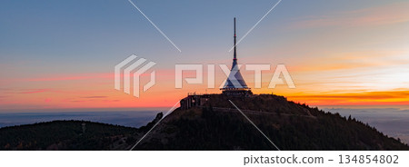 Aerial view shows Jested mountain with a hotel at sunset. The sky is filled with colors as the sun sets behind the mountain, creating a beautiful landscape in the evening. 134854802