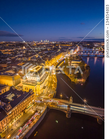 The National Theatre in Prague is lit up at night. Aerial view shows the Vltava River, Rasin embankment, and nearby architecture under the evening sky. City lights reflect on the water. 134854803