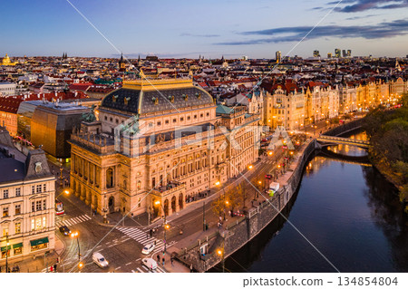 The aerial view captures the National Theatre in Prague during the evening. Lights illuminate the building as cars drive along the embankment beside the river. The aerial view captures the National Theatre in Prague during the evening. Lights illuminate the building as cars drive along the embankment beside the river. 134854804