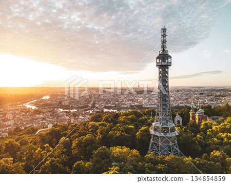 At sunrise, the Petrin Lookout Tower stands tall on a hill surrounded by lush greenery. The soft glow of morning light bathes the tower and the cityscape below in warm hues. At sunrise, the Petrin Lookout Tower stands tall on a hill surrounded by lush greenery. The soft glow of morning light bathes the tower and the cityscape below in warm hues. 134854859