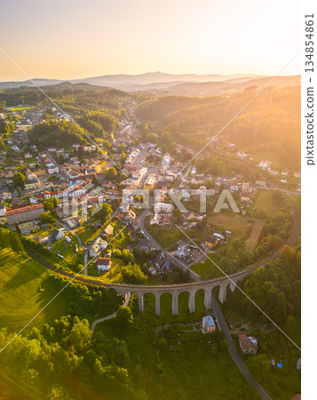 A view of a railway bridge in Smrzovka during sunset. The bridge spans across green hills and connects parts of the town. Buildings and homes nestle close together in the valley. A view of a railway bridge in Smrzovka during sunset. The bridge spans across green hills and connects parts of the town. Buildings and homes nestle close together in the valley. 134854861