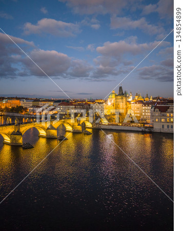 Charles Bridge stands illuminated at night over the Vltava River. The skyline of Prague features buildings and the embankment reflecting in the water. Charles Bridge stands illuminated at night over the Vltava River. The skyline of Prague features buildings and the embankment reflecting in the water. 134854899