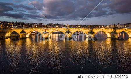 The view shows Charles Bridge in Prague during the evening. The bridge is lit up and reflects over the water below. Boats are on the river adding to the scene. 134854900