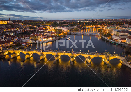 Aerial view of Charles Bridge illuminated at night. The scene showcases the water below, surrounding buildings, and the vibrant lights of Prague. Evening time highlights the beauty of this location. 134854902