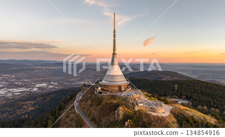 Aerial view of Jested mountain with the hotel during sunset. The sky shows warm colors as daylight fades into evening. Surrounding landscape features trees and the town of Liberec below. Aerial view of Jested mountain with the hotel during sunset. The sky shows warm colors as daylight fades into evening. Surrounding landscape features trees and the town of Liberec below. 134854916