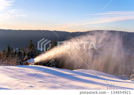 A snow cannon operates on a snowy slope in a mountain area. The device creates artificial snow against a clear blue sky. Trees line the scene, adding to the winter landscape. A snow cannon operates on a snowy slope in a mountain area. The device creates artificial snow against a clear blue sky. Trees line the scene, adding to the winter landscape. 134854921