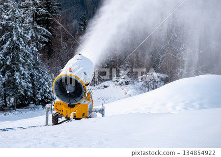 A snow cannon creates artificial snow on a winter slope. The machine blows snow into the air, covering the ground. Nearby trees are dusted with white snow in a ski area. A snow cannon creates artificial snow on a winter slope. The machine blows snow into the air, covering the ground. Nearby trees are dusted with white snow in a ski area. 134854922