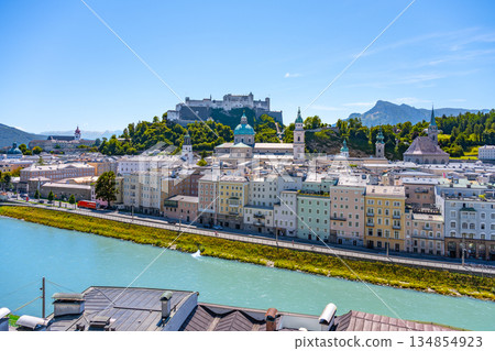 A breathtaking view of Salzburg, Austria, showcasing colorful buildings along the river with the majestic castle perched on the hill above, all under a clear blue sky. A breathtaking view of Salzburg, Austria, showcasing colorful buildings along the river with the majestic castle perched on the hill above, all under a clear blue sky. 134854923