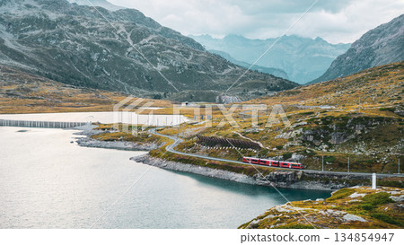 Evening light casts a serene glow over Lago Bianco at Bernina Pass. The surrounding mountains rise majestically, while a train travels along the lakeshore, enhancing the tranquil atmosphere. 134854947