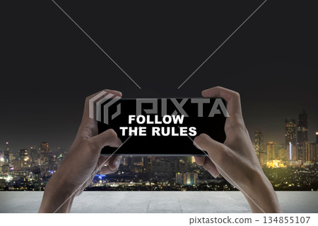 Person holding a mobile phone displaying the text message "Follow the Rules" against a cityscape at night. Concept of rules, compliance, and governance Person holding a mobile phone displaying the text message "Follow the Rules" against a cityscape at night. Concept of rules, compliance, and governance 134855107