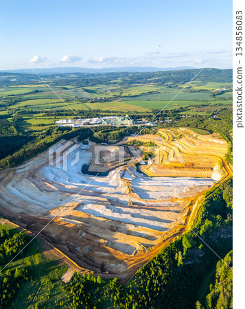 The sandstone quarry in Strelec shows terraced layers of earth. Machinery works in the distance while green hills surround the area. This site is located in Czechia's Bohemian Paradise. The sandstone quarry in Strelec shows terraced layers of earth. Machinery works in the distance while green hills surround the area. This site is located in Czechia's Bohemian Paradise. 134856083