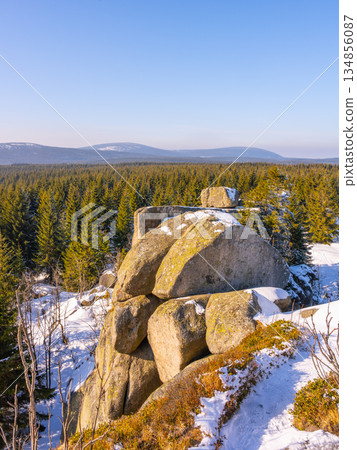 Sun shines on Pytlacke Rocks in the Jizera Mountains during a cold winter morning. Snow covers the landscape, while the serene village of Jizerka sits nearby, surrounded by trees. 134856087