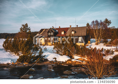 A sunny winter morning captures the tranquility of Jizerka village. Snow blankets the landscape, highlighting a charming cottage near a gentle stream. 134856090