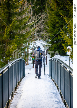 A solo skier crosses Karlov Bridge over the Jizera River on the border of Czechia and Poland during winter. The snow-covered path is surrounded by tall, green trees. A solo skier crosses Karlov Bridge over the Jizera River on the border of Czechia and Poland during winter. The snow-covered path is surrounded by tall, green trees. 134856094