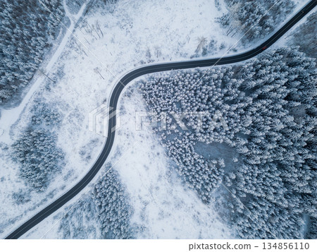 A winding road cuts through a thick forest blanketed in snow. The scene captures the serene beauty of winter in a remote area, with trees heavily covered in frost. 134856110
