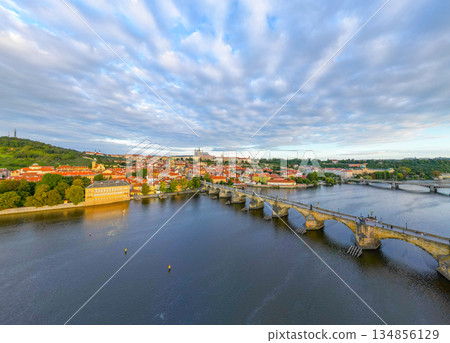 Morning light bathes the Vltava River as Charles Bridge connects old town to the iconic Prague Castle. The scene captures serene beauty with scattered clouds overhead. 134856129