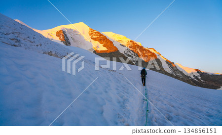 A climber ascends the snow-covered slopes of Piz Palu in the Bernina Range during summer. The stunning sunrise illuminates the surrounding glaciers and mountains. 134856151