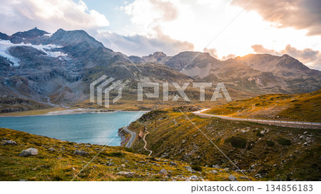 A serene evening at Lago Bianco showcases the beauty of the Swiss Alps. The sun sets behind majestic mountains, reflecting on the glacier-fed lake surface along the winding road. 134856183