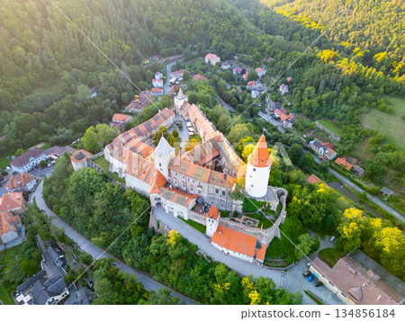 Overlooking the lush landscape, Krivoklat Castle stands majestically in Central Bohemia. The warm hues of sunset enhance its medieval charm, surrounded by serene hills and quaint villages. Overlooking the lush landscape, Krivoklat Castle stands majestically in Central Bohemia. The warm hues of sunset enhance its medieval charm, surrounded by serene hills and quaint villages. 134856184