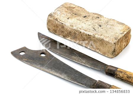 Two old, rustic knives lie next to a big thick whetstone. Blades show signs of age and wear, with wooden handles 134856833