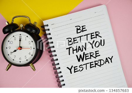 Be Better Than You Were Yesterday text written on notepad with alarm clock on pink background 134857683