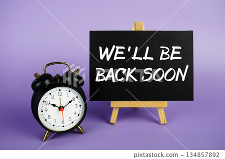 We'll Be Back Soon text with easel and alarm clock on purple background We'll Be Back Soon text with easel and alarm clock on purple background 134857892