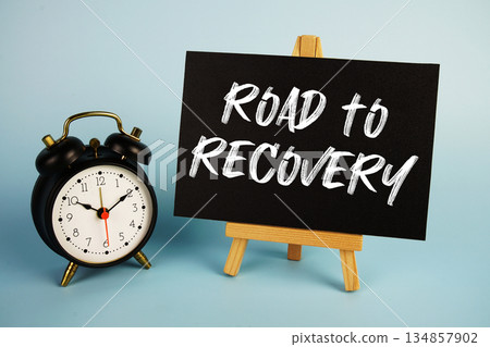 Road to Recovery text with easel and alarm clock on blue background Road to Recovery text with easel and alarm clock on blue background 134857902