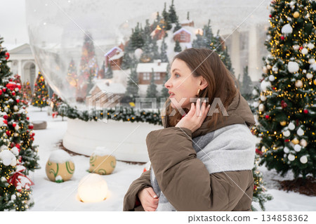 Smiling woman in warm winter jacket stands among decorated Christmas trees with red ornaments in a snowy Nutcracker-themed New Year installation Smiling woman in warm winter jacket stands among decorated Christmas trees with red ornaments in a snowy Nutcracker-themed New Year installation 134858362